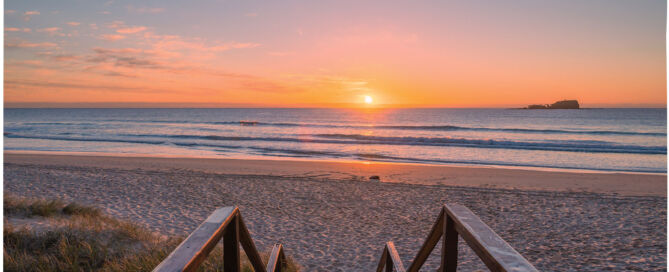 Sunrise over a sandy beach with calm ocean waves and a small island in the distance.